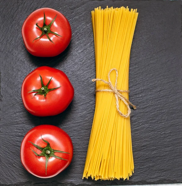 Siyah kaba bir arka planda çiğ spagetti ve kırmızı domatesler. İtalyan yemekleri pişirmek için. Yukarıdan güzel bir çiğ yemek modeli. Üç kırmızı domates ve uzun spagetti.