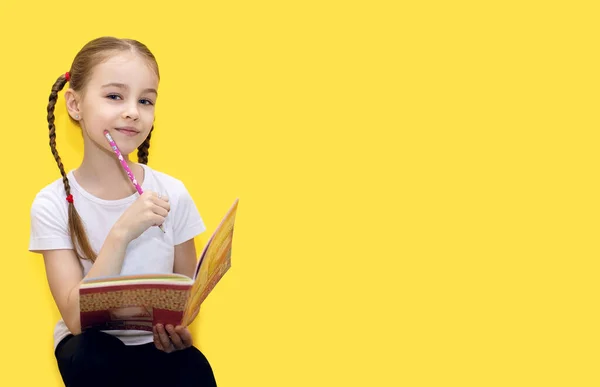 A cute girl is learning lessons holding a notebook and a pencil in her hand, sitting on a yellow background. A child with pigtails thought while doing a school assignment. Free space for text
