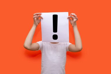 Çocuk, turuncu arka planda ünlem işareti olan bir kağıt parçası tutuyor ve yüzünü örtüyor. Güçlü duyguları ya da heyecanı ifade etme kavramı. Alarm işareti veya bir soruya yanıt