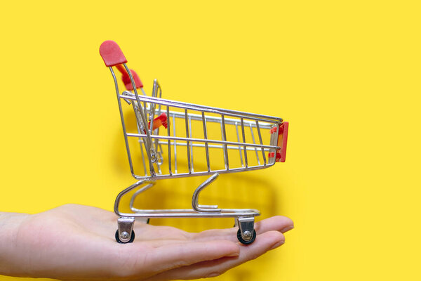 Small shopping trolley in hand on a yellow background close-up. The concept of buying and selling. Delivery of goods to the trading floors. A small toy cart with wheels. Empty shopping cart
