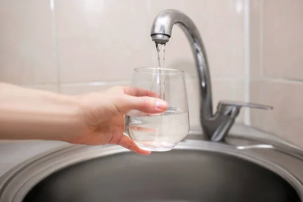A woman pours water into a transparent glass cup in the kitchen close-up. Pure fresh water from the tap at home. Benefits for the human body from drinking water