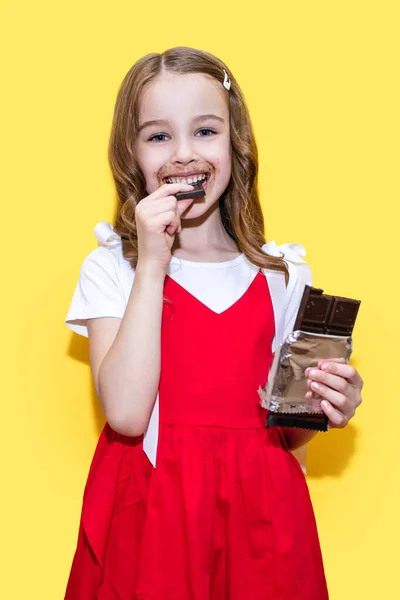 Κορίτσι Τρώει Μια Σοκολάτα Κόκκινο Φόρεμα Κίτρινο Φόντο Αστείο Παιδί — Φωτογραφία Αρχείου