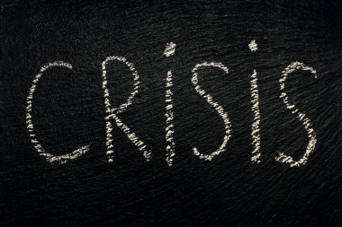 The word crisis written in white chalk on a black rough surface. Difficult situation that needs to be addressed. Writing text with chalk on a black board