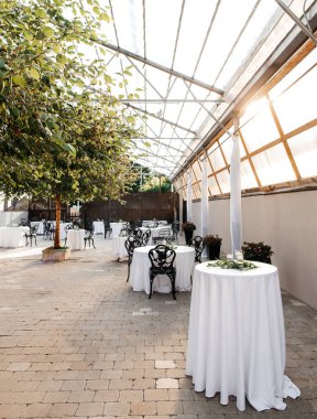 A restaurant with a transparent roof with decorated tables and trees in the center. Preparation for the reception of guests and table setting. Restaurant with transparent roof and beautiful decor