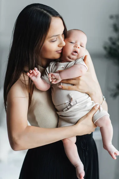 A young mother holds her newborn son in her arms in a bright bedroom. Happy mother of a toddler in a vertical photo. Portrait of happy mother and child in apartment