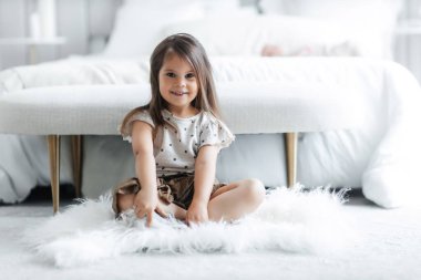 A cute cheerful girl sits on the floor in a bright room and smiles. Happy child with big eyes on the background of the bed. Free space for text