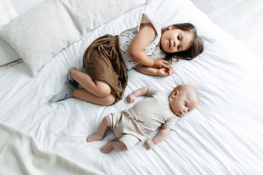 Little girl and her newborn brother are lying on the bed in the bedroom, top view. The children are resting on the bed. The children fall asleep in their bedroom