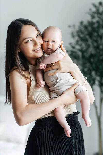 A young mother holds her newborn son in her arms in a bright bedroom and smiles. Happy mother of a toddler in a vertical photo. Portrait of happy mother and child in apartment