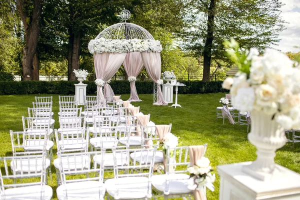 Beautiful wedding arch and white chairs decorated with white flowers on a green lawn. Preparing for the wedding ceremony. The arch for the wedding ceremony and chairs for guests are decorated