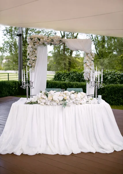 The wedding banquet table of the newlyweds is decorated with an arch of flowers and high candlesticks with candles. Beautifully decorated table of the bride and groom with flowers