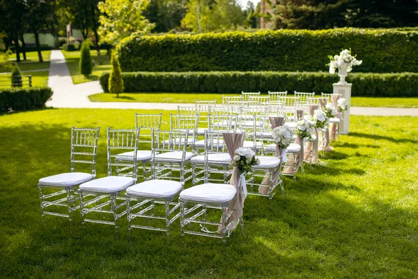 White chairs on a green lawn for a wedding ceremony are waiting for guests. Beautifully decorated seating areas in the form of chairs with flowers and fabrics. Preparing for the wedding ceremony