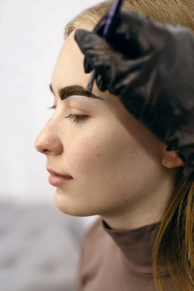 Eyebrow master shapes and paints with a brush the eyebrows of a red-haired girl close-up in a beauty salon. Young woman in the process of dyeing her eyebrows on a vertical background