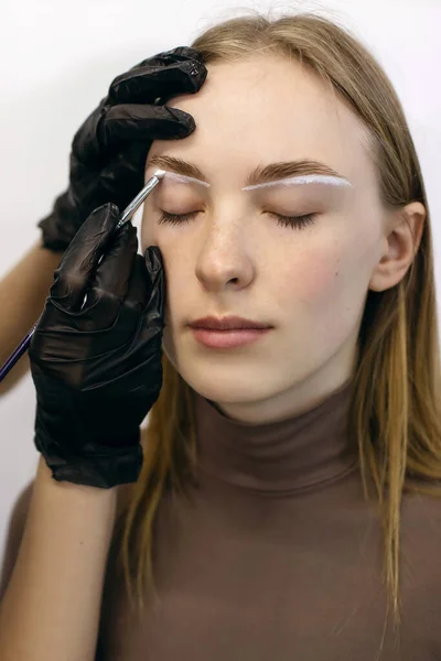 Red-haired girl with freckles in a beauty salon makes eyebrows. The eyebrow master draws a white outline around the eyebrows of a freckled young woman for further coloring