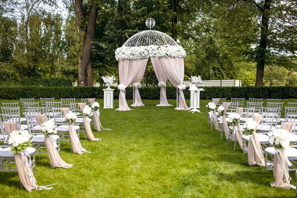 Beautiful wedding arch and white chairs on a green lawn for a wedding ceremony. The arch for the wedding ceremony of the newlyweds and the chairs for the guests are decorated with white roses