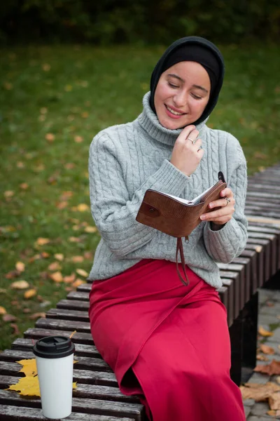 グレーのセーターを着た若いイスラム教徒の女性がベンチにノートを置いて座っています コーヒーと秋の葉のカップ 学生だ 大学生 — ストック写真