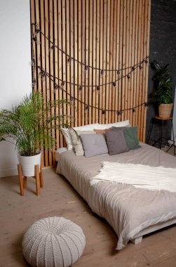 Yatak odası. Gri çarşaflı yatak, yastıklar. Arka planda Areca chrysalidocarpus çiçeği var. Tahta duvar ve çelenk duvarda. Bir çocuk için küçük bir puf ve sallanan bir at..