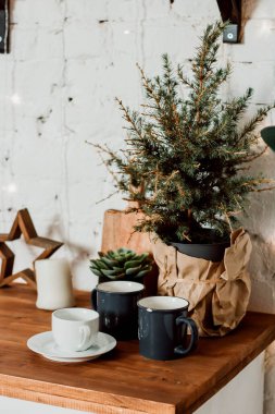 Mutfak setindeki bir tencerede küçük bir Noel ağacı. Bardaklar, fincan ve çay tabağı masanın üstünde..