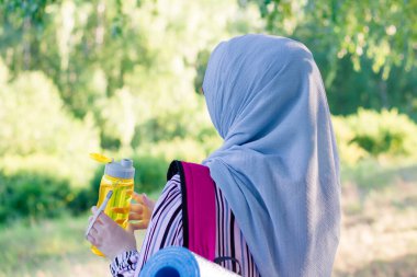 Sırt çantalı Müslüman bir kadın ve su için sarı bir şişeyle ormanda yürüyüş paspası. Ormanda yürüyüş. Seyahat. Pembe sırt çantası..