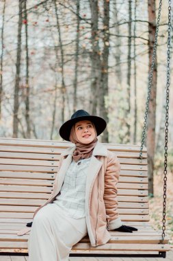 Bej ceketli ve türbanlı Müslüman bir kadın. Siyah şapka. Parktaki şık Müslüman kadın. Salıncakta oturan bir kız