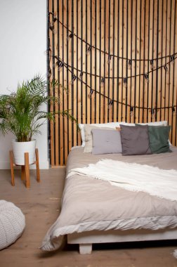 Yatak odası. Gri çarşaflı yatak, yastıklar. Arka planda Areca chrysalidocarpus çiçeği var. Tahta duvar ve çelenk duvarda. Bir çocuk için küçük bir puf ve sallanan bir at..