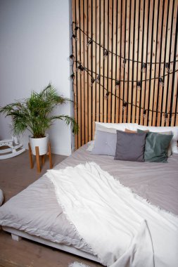 Yatak odası. Gri çarşaflı yatak, yastıklar. Arka planda Areca chrysalidocarpus çiçeği var. Tahta duvar ve çelenk duvarda.