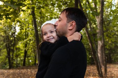 Baba ve kızı sarılıyorlar ve kız kameraya bakıyor. Yaz parkı. Yeşil ağaçlar. Genç aile kavramı. Yıldızlı süsleme kızın saçına örülmüş.. 