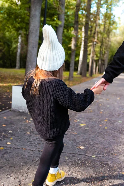 Mom and daughter. Mom is holding her daughter\'s hand. A girl in a black sweater and a white hat. Tenderness of mother and daughter. Summer park. Green trees. The concept of a young family.