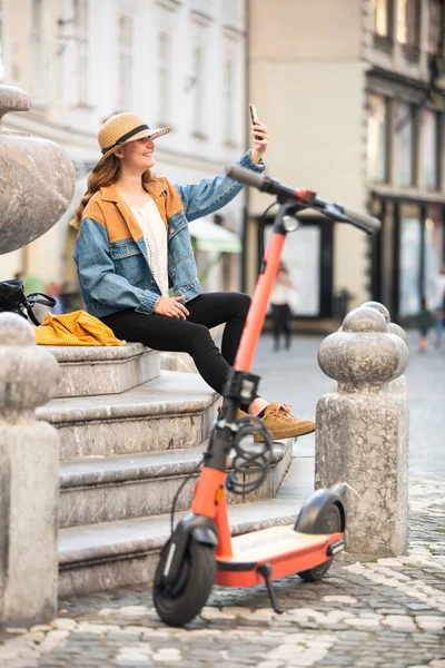 Молодая женщина, сидя на лестнице старинного исторического здания в центре города, фотографирует на телефон. Путешествующая женщина исследует старый город Любляны на электрическом скутере. Летний отдых. — стоковое фото