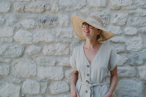 Retrato de una hermosa mujer rubia alegre con un vestido de verano de una pieza y un sombrero de verano, de pie frente a la antigua pared de piedra medieval. concepto de retrato de vacaciones de verano. —  Fotos de Stock