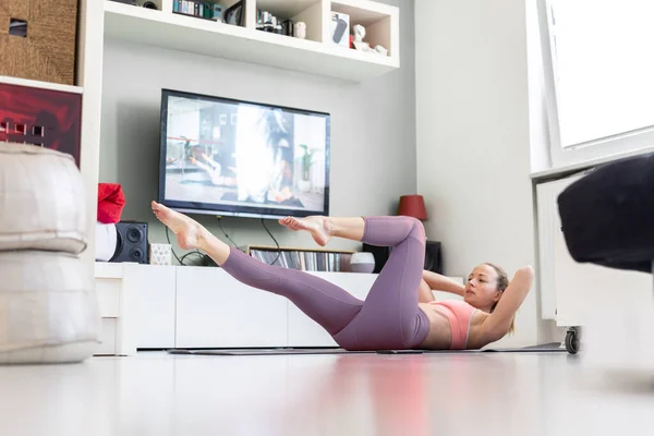 Atractiva mujer deportiva haciendo ejercicio en casa, haciendo ejercicios de pilates frente a la televisión en su pequeño apartamento de estudio. Distanciamiento social. — Foto de Stock