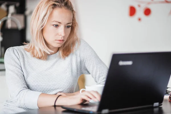 Kvinnlig frilansare i vardagskläder som arbetar på distans från matbordet på morgonen. Hem kök i bakgrunden. — Stockfoto