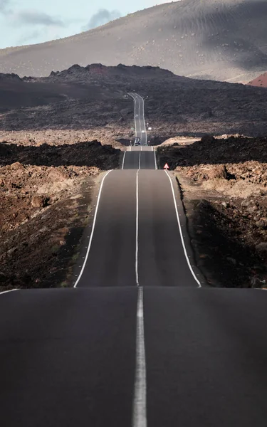 Endless road on a volcano in Timanfaya National Park in Lanzarote in the Canary Islands with a continuous line, black volcanic rocks on the side and volcanoes in mist in background. — Stock Photo, Image