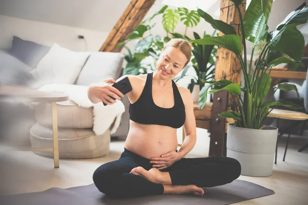 Young happy and cheerful beautiful pregnant woman taking selfie with her mobile phone while staying fit, sporty and active on her maternity leave. Motherhood, pregnancy, yoga concept.