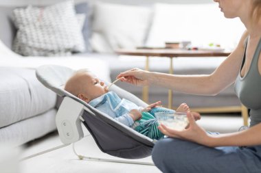 Anne, bebeğini bebek koltuğunda meyve püresiyle besliyor. Bebek katı gıda tanıtım konsepti