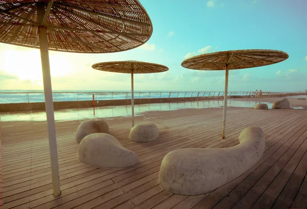 Wooden deck with stone benches and metal with straw umbrellas — Stock Photo, Image