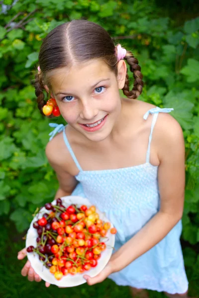 Красивая маленькая улыбающаяся девочка ест вишни в саду — стоковое фото