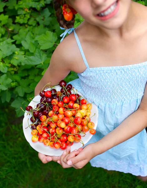 Красивая маленькая улыбающаяся девочка ест вишни в саду — стоковое фото