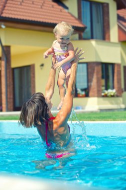 Summer vacation. Young woman plaing with her little daughter in swimming pool outdoors clipart