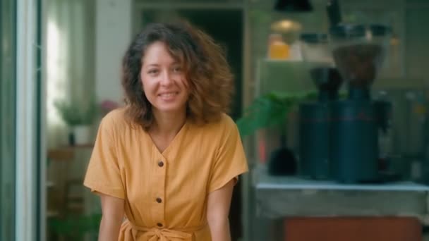 Sorrindo mulher encaracolado em vestido amarelo mostrando língua dançando feliz enquanto se senta no café — Vídeo de Stock