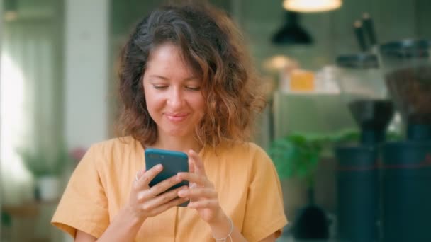 Χαμογελώντας σγουρή γυναίκα σε κίτρινο φόρεμα χρησιμοποιώντας το τηλέφωνό της ευτυχώς, ενώ κάθεται στην καφετέρια — Αρχείο Βίντεο