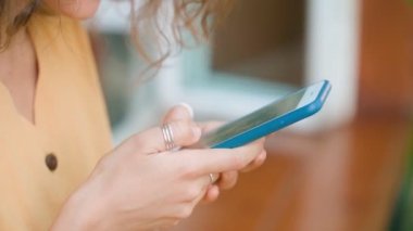 Sarı elbiseli, gülümseyen kıvırcık kadın kahvecide otururken telefonunu mutlu bir şekilde kullanıyor.