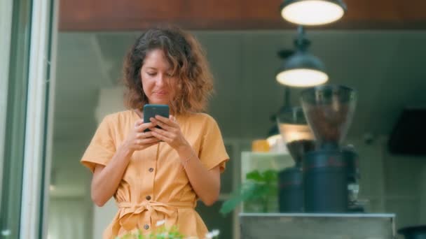 Uśmiechnięta kręcona kobieta w żółtej sukience korzystająca ze swojego telefonu szczęśliwie siedząc w kawiarni — Wideo stockowe