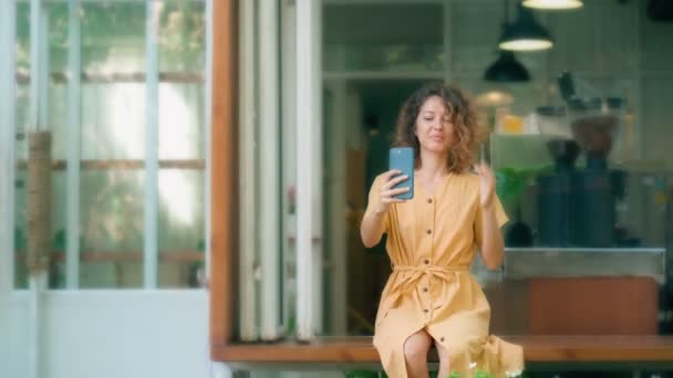 Glimlachende krullende vrouw in gele jurk met behulp van haar telefoon gelukkig terwijl zit in de coffeeshop — Stockvideo