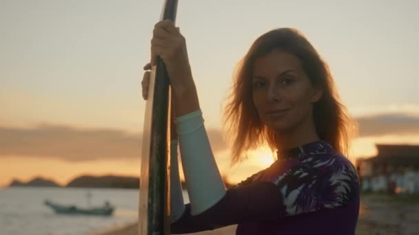 Jonge aantrekkelijke vrouwelijke surfer glimlachend en poserend met haar surfplank bij zonsondergang op het strand — Stockvideo