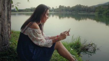 Transseksüel Asyalı kadın parkta bir ağacın yanında oturuyor, telefonuyla mesajlaşıyor..