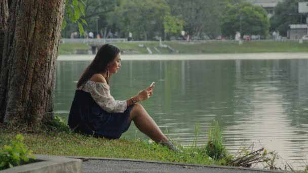 Transznemű ázsiai nő ül egy fa mellett a parkban, SMS-ezik a telefonján.. — Stock videók