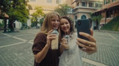 Two young smiling girls take selfie photos on a smartphone. Models pose in front of the street.