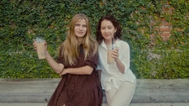Two women sit on a bench near a wall of vines and drink drinks — Stock video