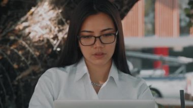 Asyalı çekici, gözlüklü genç bir kadın, dışarıda bir teras kafede laptopunun başında çalışıyor.
