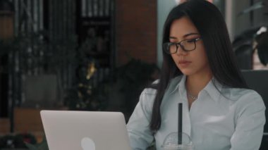 Asyalı çekici, gözlüklü genç bir kadın, dışarıda bir teras kafede laptopunun başında çalışıyor.